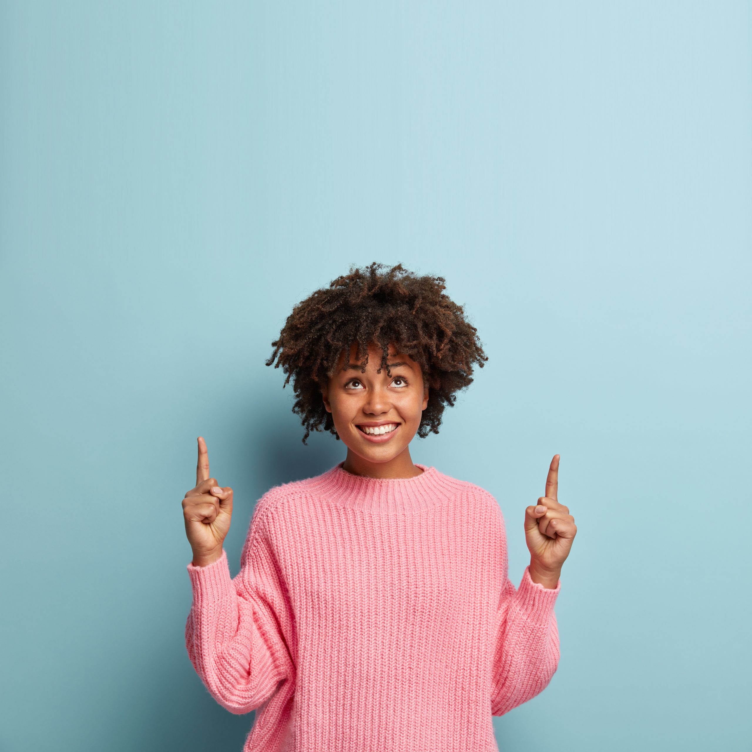 Vertical shot of good looking smiling lady with Afro haircut, wears loose pink sweater, shows direction upwards, poses over light blue background, demonstrates blank space for your advertisement